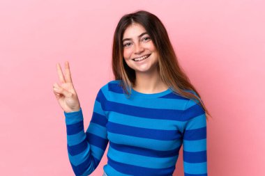 Pembe arka planda izole edilmiş genç beyaz kadın gülümsüyor ve zafer işareti gösteriyor.