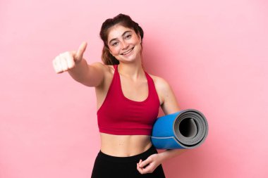 Genç sporcu kadın, elinde minderle yoga derslerine gidiyor ve baş parmak hareketi yapıyor.