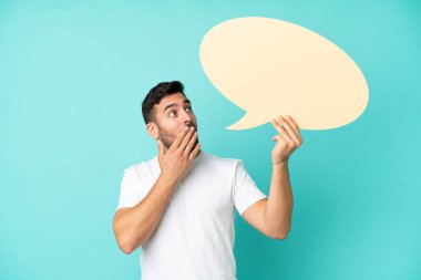 Mavi arka planda izole edilmiş yakışıklı beyaz bir adam elinde şaşırmış bir ifadeyle boş bir konuşma balonu tutuyor.