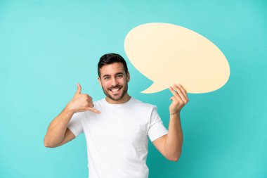 Mavi arka planda izole edilmiş yakışıklı beyaz bir adam elinde boş bir konuşma balonu ve telefon jesti yapıyor.