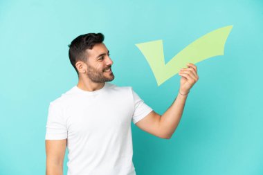Mavi arka planda izole edilmiş yakışıklı beyaz bir adam elinde mutlu bir ifadeyle bir çek simgesi tutuyor.