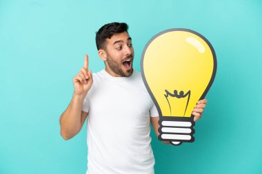 Mavi arka planda izole edilmiş yakışıklı beyaz adam elinde bir ampul simgesi tutuyor ve düşünüyor.