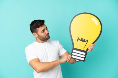 Mavi arka planda izole edilmiş yakışıklı beyaz bir adam elinde bir ampul simgesi ve şüpheleri var.