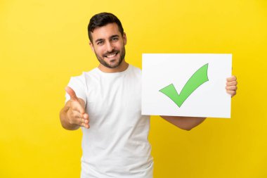 Sarı arka planda izole edilmiş yakışıklı beyaz bir adam. Elinde yeşil işaret simgesi olan bir pankart var. Anlaşma yapıyor.
