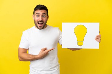 Sarı arka planda izole edilmiş yakışıklı beyaz bir adam elinde ampul simgesi olan bir pankart tutuyor ve onu işaret ediyor.