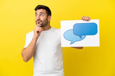 Sarı arka planda izole edilmiş yakışıklı beyaz bir adam konuşma balonu ikonlu bir levha tutuyor ve düşünüyor.