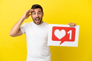 Young handsome caucasian man isolated on yellow background holding a placard with Like icon with surprised expression