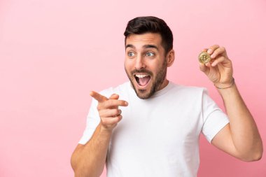 Elinde pembe arka planda izole edilmiş bir Bitcoin tutan genç adam, parmağını yana uzattı ve bir ürün sundu.