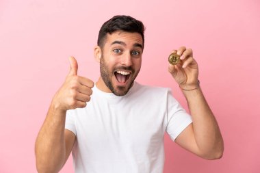 Elinde pembe arka planda izole edilmiş bir Bitcoin tutan genç adam. Başparmakları havada çünkü iyi bir şey oldu.