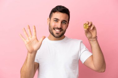 Elinde pembe arka planda izole edilmiş Bitcoin tutan genç adam parmaklarıyla beşi sayıyor.