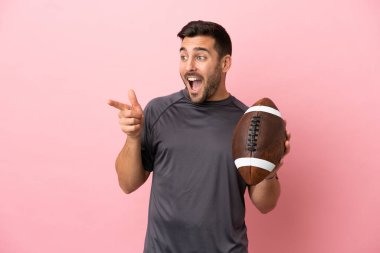 Rugby oynayan beyaz bir adam pembe arka planda izole edilmiş bir şekilde parmak kaldırıyor ve bir ürün sunuyor.