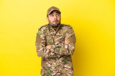 Military man isolated on yellow background making doubts gesture while lifting the shoulders