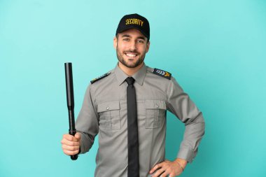 Mavi arka planda izole edilmiş genç güvenlik görevlisi kolları kalçasında ve gülümsüyor.