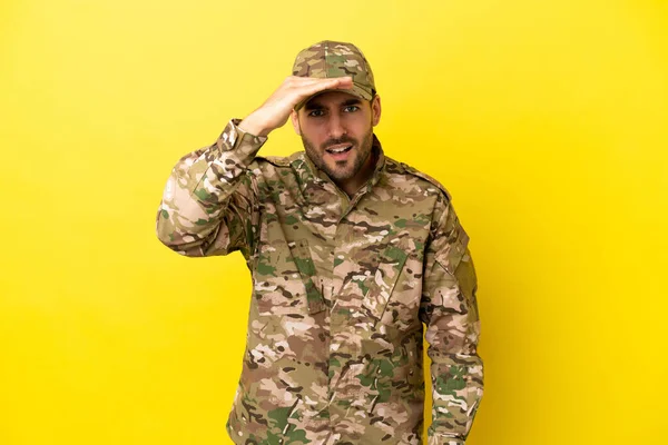 Military man isolated on yellow background looking far away with hand to look something