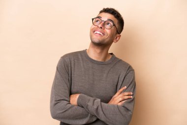 Genç Brezilyalı adam bej arka planda izole edilmiş gülümserken yukarı bakıyor