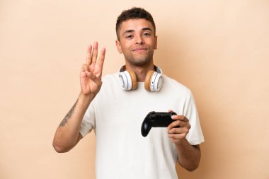 Bej arka planda izole edilmiş bir bilgisayar oyunu kontrolörüyle oynayan genç Brezilyalı adam mutlu ve parmaklarıyla üçe kadar sayıyor.