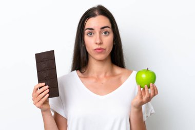 Beyaz arka planda izole edilmiş beyaz bir kadın bir elinde çikolata tableti, diğerinde elma alıyor.