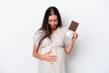 Beyaz arka planda izole edilmiş beyaz bir kadın hamile ve elinde sürpriz bir ifadeyle çikolata tutuyor.