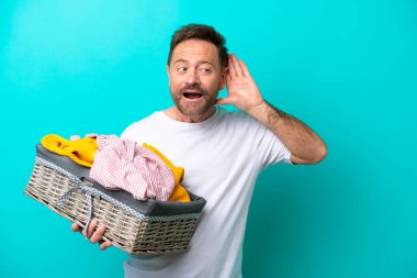 Orta yaşlı bir kadın mavi arka planda izole edilmiş bir çamaşır sepetini tutuyor. Kulağına elini sürerek bir şey dinliyor.