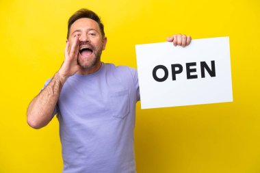 Orta yaşlı beyaz bir adam sarı arka planda izole edilmiş. Elinde açık yazılı bir pankart var ve bağırıyor.