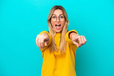Mavi arka planda izole edilmiş genç Uruguaylı kadın şaşırmış ve ön tarafı işaret ediyor.