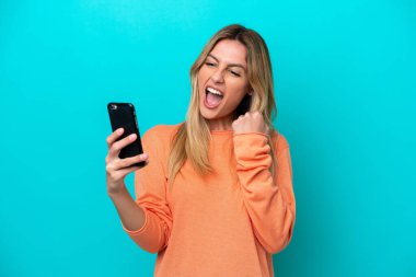 Genç Uruguaylı kadın mavi arka planda izole edilmiş ve telefon zafer pozisyonunda.