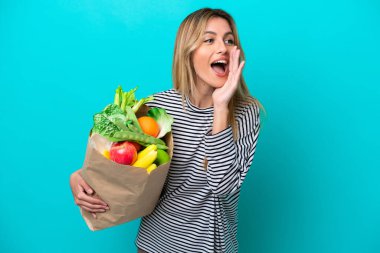 Genç Uruguaylı kadın elinde alışveriş poşeti tutarken mavi arka planda izole bir şekilde ağzı sonuna kadar açık bağırıyor.