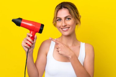 Genç Uruguaylı kadın elinde saç kurutma makinesi ile Don sarı arka planı izole ediyor ve onu işaret ediyor.