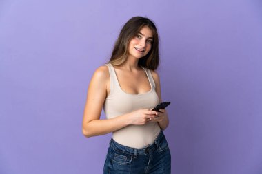 Mor arka planda izole edilmiş genç beyaz kadın cep telefonuyla bir mesaj gönderiyor.