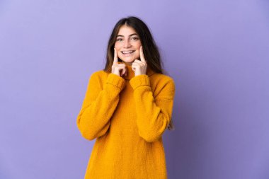 Mor arka planda izole edilmiş genç beyaz kadın mutlu ve hoş bir ifadeyle gülümsüyor.