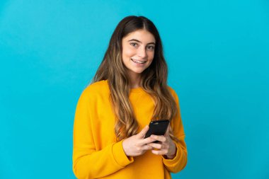 Mavi arka planda izole edilmiş genç beyaz kadın kameraya bakıyor ve cep telefonunu kullanırken gülümsüyor.