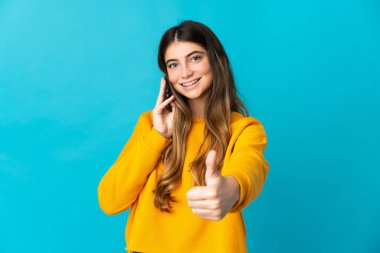 Mavi arka planda izole edilmiş genç beyaz kadın başparmaklarını kaldırırken cep telefonuyla sohbet ediyor.