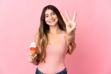 Kornet dondurmalı genç beyaz kadın pembe arka planda izole edilmiş mutlu ve parmaklarıyla üçe kadar sayıyor.