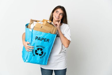 Elinde kağıt dolu bir geri dönüşüm çantası tutan genç bir kadın beyaz arka planda izole edilmiş bir şekilde bir fikri düşünürken yukarı bakıyor.