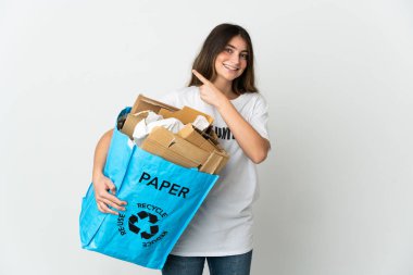 Elinde kağıt dolu bir geri dönüşüm çantası tutan genç bir kadın beyaz arka planda izole edilmiş bir şekilde bir ürünü sunmak için kenarı işaret ediyor.