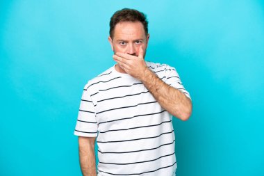 Orta yaşlı, beyaz bir adam, mavi arka planda izole edilmiş, ağzı elle kapatılmış.