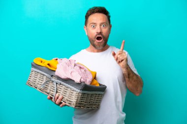 Orta yaşlı bir kadın mavi arka planda izole edilmiş bir çamaşır sepetini tutarken parmağını kaldırarak çözümü bulmayı umuyor.