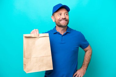 Orta yaşlı bir adam mavi arka planda izole edilmiş bir çanta dolusu yiyecek alıyor. Kolları kalçasında ve gülümseyerek poz veriyor.
