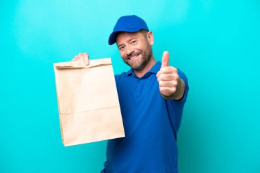 Orta yaşlı bir adam mavi arka planda izole edilmiş bir paket yiyecek alıyor. Başparmakları havada çünkü iyi bir şey oldu.