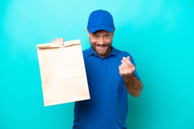 Orta yaşlı bir adam mavi arka planda izole edilmiş bir paket yiyecek alıyor ve para hareketi yapıyor.