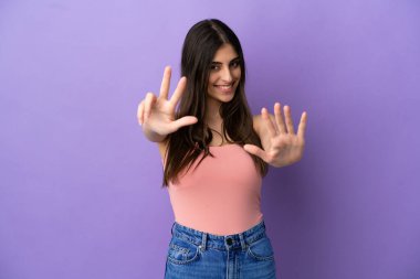 Mor arka planda izole edilmiş genç beyaz kadın parmaklarıyla sekiz sayıyor.