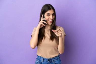 Mor arka planda izole edilmiş cep telefonu kullanan beyaz bir kadın şaşırmış ve ön tarafı işaret ediyor.