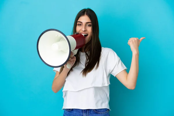 Young caucasian woman isolated on blue background shouting through a megaphone and pointing side