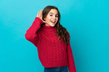 Mavi arka planda izole edilmiş küçük beyaz kız elini kulağına koyarak bir şeyler dinliyor.