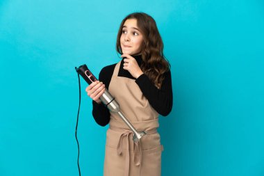 Mavi arka planda izole edilmiş el blenderi kullanan küçük bir kızın şüpheleri ve düşünceleri var.