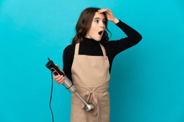 Mavi arka planda el blenderi kullanan küçük kız yan tarafa bakarken sürpriz jest yapıyor.