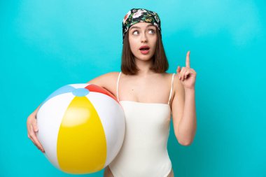 Elinde plaj topu tutan genç Ukraynalı kadın mavi arka planda izole edilmiş bir şekilde parmağı yukarı kaldıran bir fikir düşünüyor.