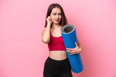 Genç sporcu Ukraynalı kadın yoga derslerine giderken elinde pembe arka planda izole edilmiş bir minder tutarken hayal kırıklığına uğramış ve kulaklarını kapatmış.