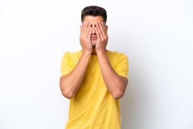 Beyaz arka planda izole edilmiş beyaz tenli genç adam gözlerini elleriyle kapatıyor.