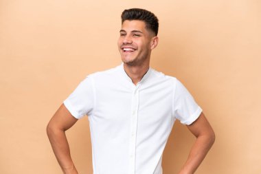 Beyazlı genç bir adam bej arka planda izole edilmiş. Kolları kalçasında ve gülümsüyor.
